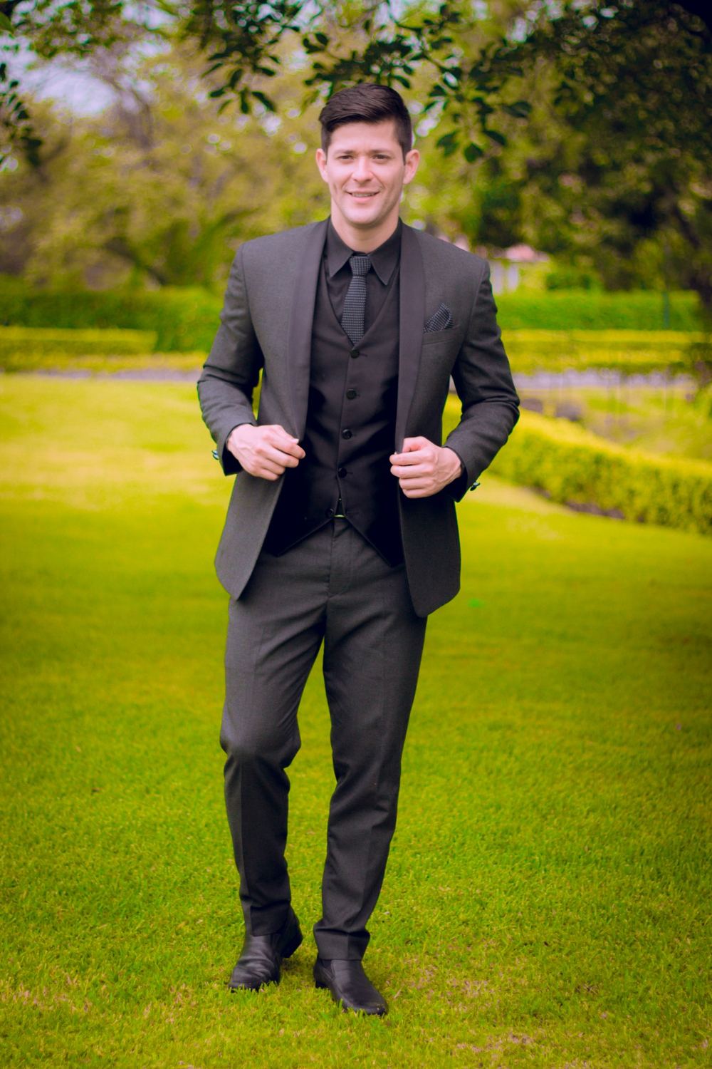 Caballero con traje gris oscuro de tres piezas y corbata a cuadros posando en jardín