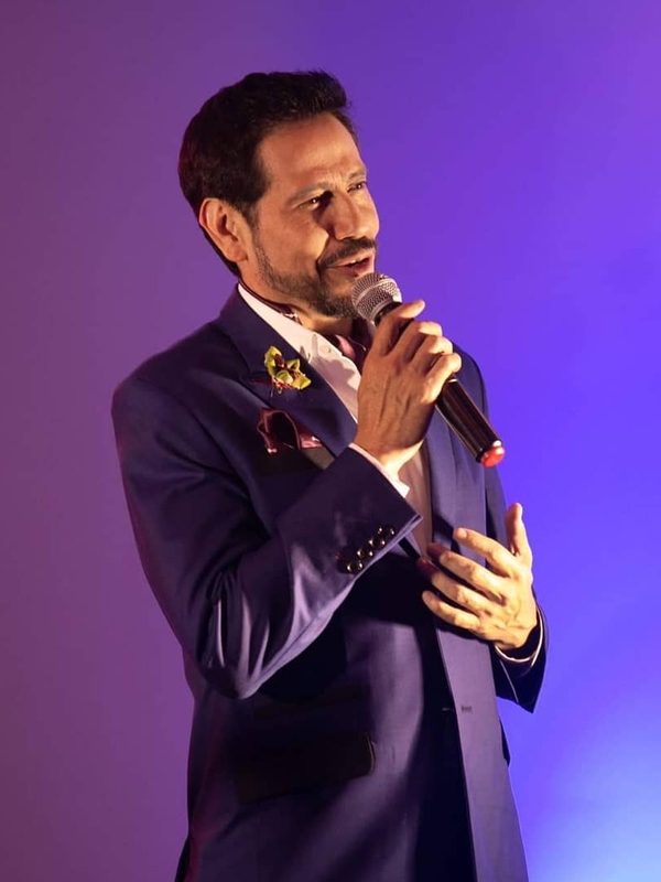 Caballero con traje azul marino y corbatín hablando en un evento con micrófono