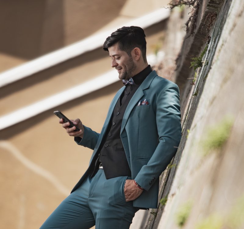 Caballero con traje verde azulado de tres piezas y corbatín floral revisando el celular